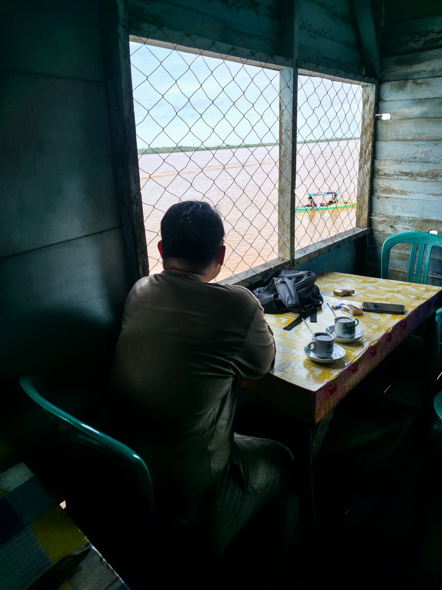 Foto di atas menangkap suasana seorang pria yang duduk sendiri, memandang jauh ke arah sungai dari balik jendela, seolah tenggelam dalam pikirannya. Ia ditemani secangkir kopi, mungkin sedang merenung, atau barangkali sedang memikirkan percakapan yang baru saja terjadi. Ada keheningan, ada jeda untuk mendengarkan—sebuah momen yang sering kita lupakan dalam kehidupan sehari-hari.